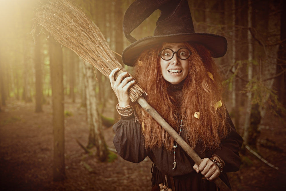 Witch with unruly red hair holding a broom in a forest, illustrating hair habits to ghost this Halloween.
